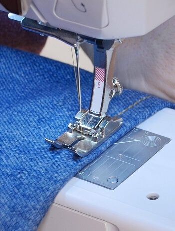 Most people try to avoid denim fabric because it is thick, meaning that its thicker seams are much harder to work with. However, sewing denim is not that difficult if done correctly. Before even diving into the best way of sewing this material, you should have a sewing machine with a powerful motor so that it can puncture through the thick layers easily. The machine should also have a high presser foot lift so that these thick seams can fit under it with ease. Another desirable feature would be a large working table so that you can level the denim fabric for a more comfortable sewing experience. With such a sewing machine in place, below are some of the tips that can help you sew the perfect denim stitches. Steam and heat Being a thick and tough material, denim might bend and form ripples after stitching, which is not a good result. The key to avoiding this outcome is to steam and heat the fabric using a steam iron to soften the fabric and make it “loosen up” a little bit. Apart from preventing these ripples, the softening also makes it easier to puncture through, giving the sewing machine an easier time doing its job. Use heavy-duty needles (specifically meant for denim) Apart from using a machine with a strong motor, you need heavy-duty needles for this job. There is a big difference between all-purpose needles and needles marked with a “jeans” tag. Jeans needles are much thicker, and therefore ideal for puncturing through heavy fabrics. The smaller 90/14 size needles can work on light denim, but heavy denim requires 100/16 or 100/18 sized needles. Use heavy-duty thread This type of thread is particularly good for topstitching, which reinforces the seams, enabling them to handle stress easily. Thick, heavy-duty threads also have a decorative effect when used for topstitching and, thus, also give the denim joints a good look. Hold it firmly when sewing This is why you need a machine with a large working table. Sometimes denim might stretch or compact if not held firmly. While sewing, you need to hold it firmly both on the front side and backside of the needle. A large working table would be ideal to do this because you will have enough space in front of and behind the needle to place your hands on the fabric as you guide through the presser foot. Use a longer stitch length Being quite tough, denim can still hold itself together when sewn using long stitches. The recommended stitch length is about 3mm, and this gives you just enough space to hold enough denim fibers per stitch. A longer stitch length also reduces strain on the needle and machine motor because they puncture fewer holes per given length. Go slow When you encounter these thick seams, it might be necessary to hand crank the wheel at times so that you do not damage the motor as well as the needles. Conclusion Even as you strive to make these durable stitches, your denim design will still be weak if it is not finished off by strong closures. Heavy-duty zippers, snaps, and buttons should be used so that the entire final design is durable and strong enough to handle a reasonable amount of stress. Related Posts: How to Sew a Bound Seam with a Sewing Machine How To Sew Leather On A Home Sewing Machine How To Sew A Button With A Sewing Machine
